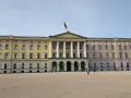 Det gula **Oslo slott** med sin flagga reser sig stolt mot en klarblå himmel. En stor öppen plats framför slottet.