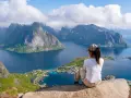 En person sitter på en klippa och blickar ut över en dramatisk kustlinje med berg och fjordar. En perfekt vy för lofoten vandring.