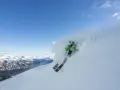 En skidåkare i grön jacka plöjer genom pudersnö med en fantastisk fjordvy i bakgrunden. Stranda ski-skidorna skär genom snön.
