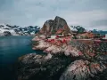 Röda fiskestugor vid en klippig kust i **Hadeland, Norge**, med snöklädda berg i bakgrunden.