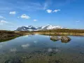 En klarblå himmel med moln speglar sig i en lugn sjö. I bakgrunden reser sig snöklädda berg, en vacker vy för ramundberget vandring.