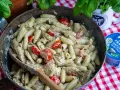 En stor panna med krämig pasta pesto, körsbärstomater och kyckling. En träsked ligger i pannan.