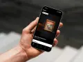 En hand håller en mobil med en app för **Nasjonalmuseet Oslo**. Appen visar en rundtur med "Skriet" och alternativ för utställningar.