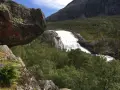 En mäktig fors forsar nerför bergssluttningen, omgiven av grönskande skog och stenblock. En perfekt vy för vandring på Hardangervidda.