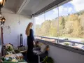 En kvinna njuter av utsikten från en liten balkong med glaspartier. Träd och en parkering syns utanför.