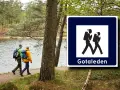 Två vandrare med ryggsäckar följer stigen längs en sjö. Skylt visar vandringsled Gotaleden etapp 6.