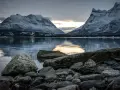 Vinterlandskap med snöklädda berg i Lyngenalperna, speglade i ett lugnt vatten. Stenar ligger vid strandkanten.