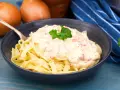 En skål med tagliatelle täckt av krämig ost och skinksås, garnerad med persilja. Två gula lökar syns i bakgrunden.
