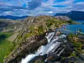 En person på en hängbro över ett vattenfall under en vandringsresa i Norge. Dramatiskt landskap med berg och en blå sjö.