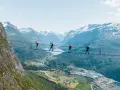 Fyra personer går på en hängbro vid Loen via ferrata, med en fantastisk utsikt över fjord och snöklädda berg.