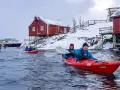 Glada paddlare i kajak i Lofoten, med snöklädda röda hus och ett stort fartyg i bakgrunden.