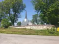 Kyrka med spira bakom stenmur och träd. En skylt visar "2 St". Kanske en vy från Hamar, Norge.