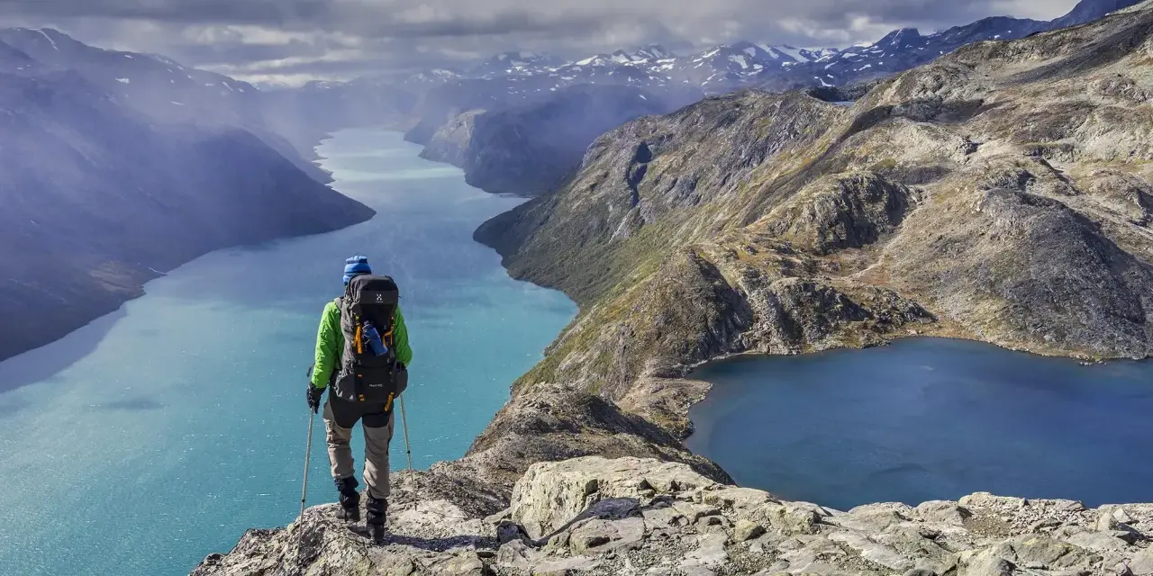 Vandrare med ryggsäck står på en klippa och blickar ut över turkosa sjöar och berg i Jotunheimen nationalpark.
