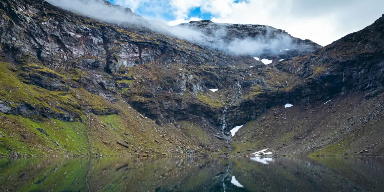 En spegelblank sjö reflekterar en dramatisk bergssida med en forsande vattenfall. Perfekt för att vandra i Abisko.