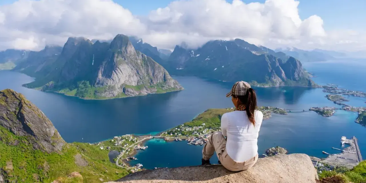 En person sitter på en klippa och blickar ut över en dramatisk kustlinje med berg och fjordar. En perfekt vy för lofoten vandring.