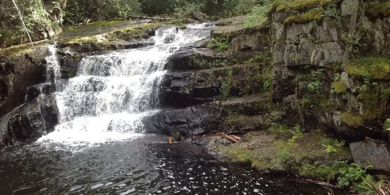 Vattenfall i en skogsmiljö nära Lillehammer, Norge. Mossbeklädda klippor och mörkt vatten.