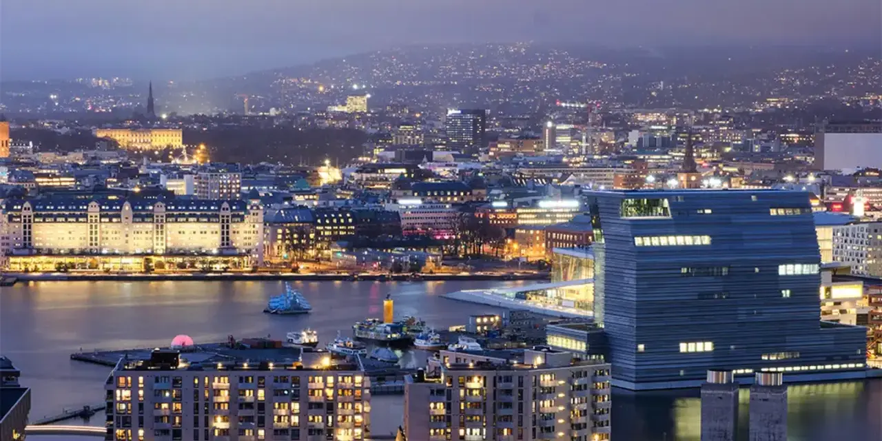 Fakta om Oslo: En stad vid vattnet med moderna byggnader och upplysta hus, som sträcker sig mot en kulle i fjärran.