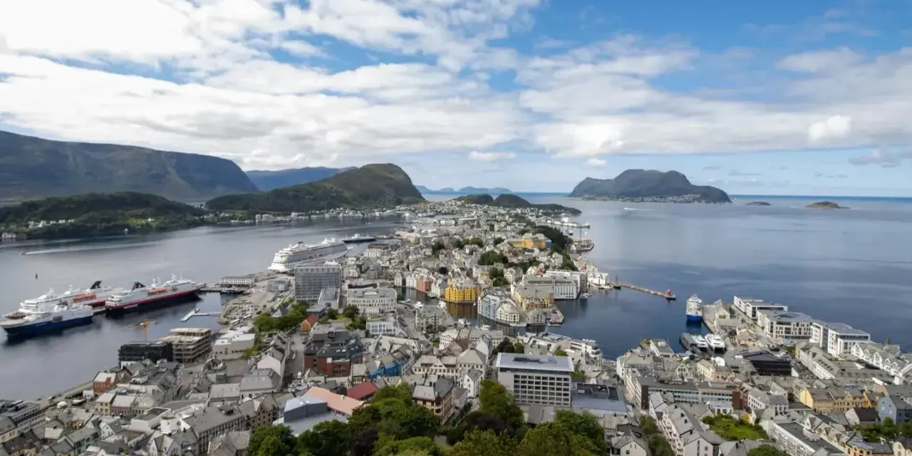 Vacker vy över Ålesund, Norge, med färgglada hus, fartyg i hamnen och berg i bakgrunden under en molnig himmel.