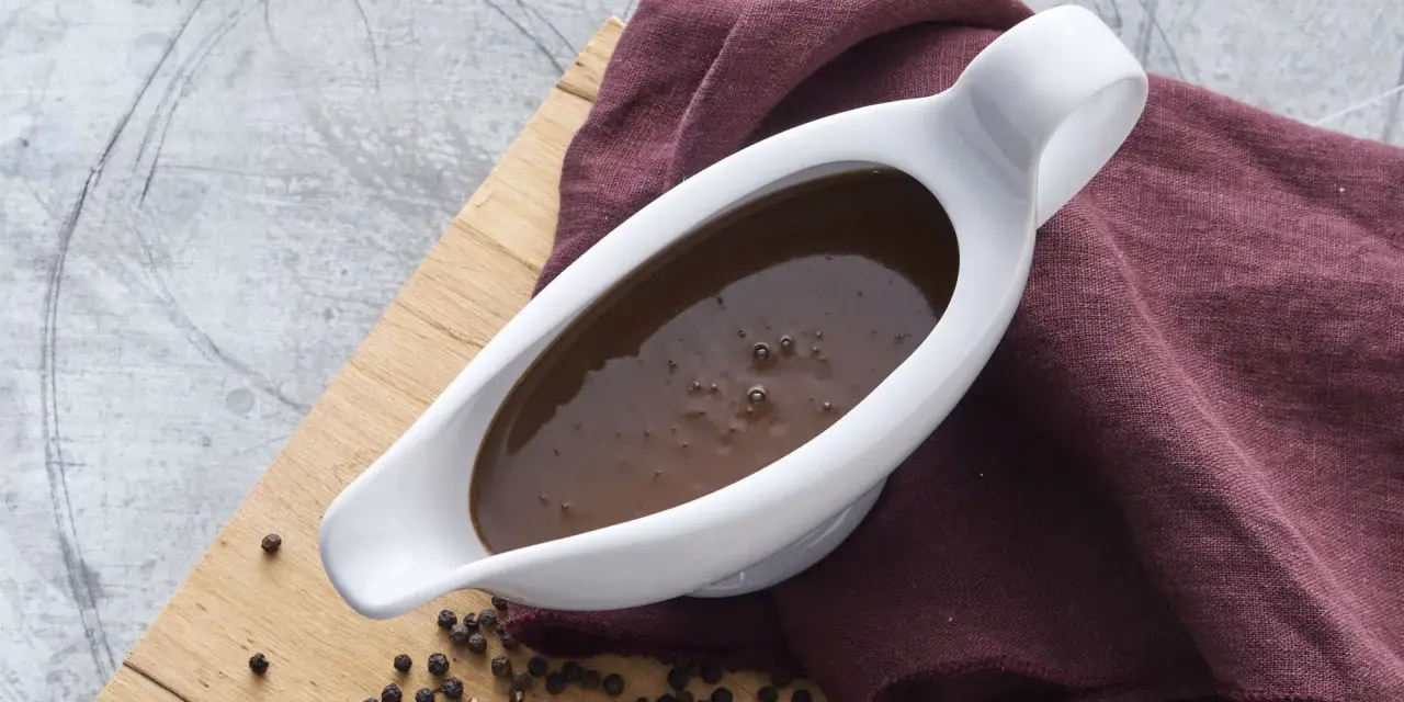 En vit såsslev fylld med mustig brunsås, bredvid en hög med svartpepparkorn. Ett perfekt brunsås recept för middagen.