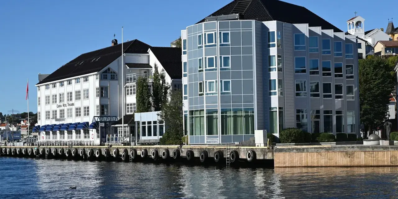 Hotellbygg vid vattnet i Arendal, Norge. En vit byggnad med blå markiser och en modern glasbyggnad står sida vid sida.