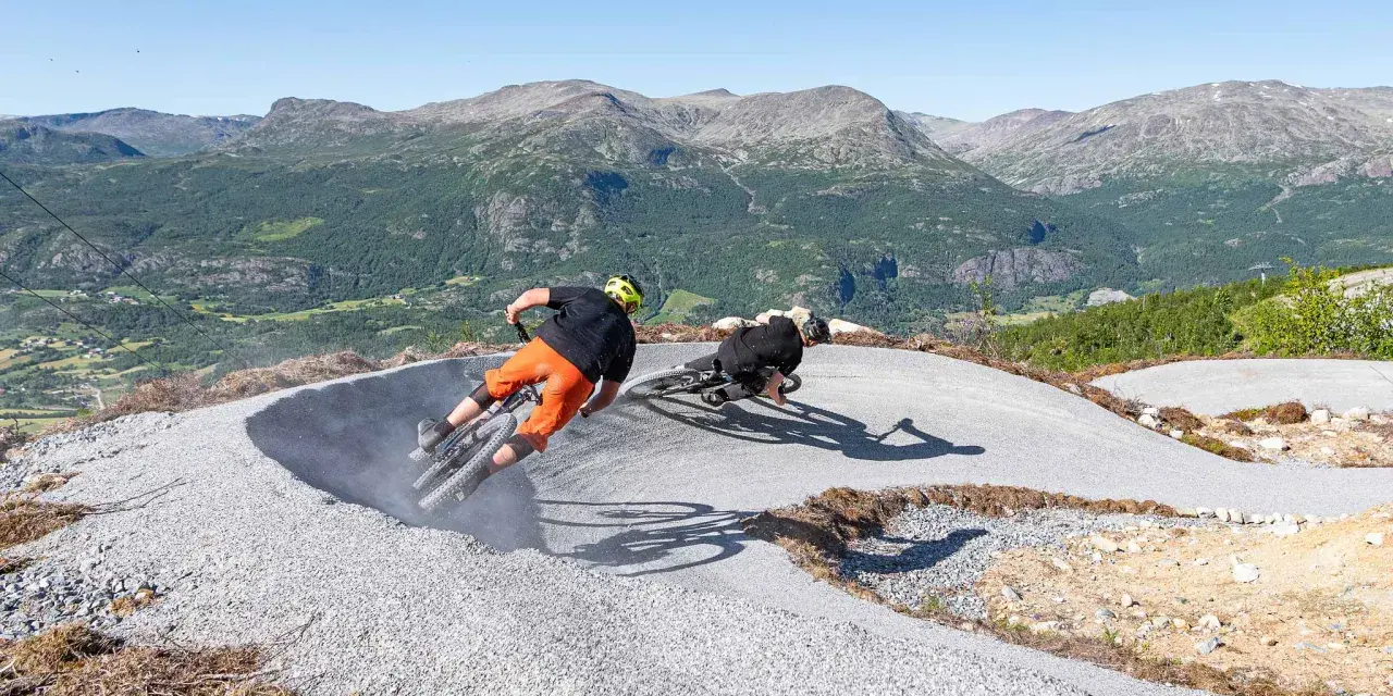 Två cyklister kör utför en grusig bana i Hemsedal sommar. Berg och grönska i bakgrunden.