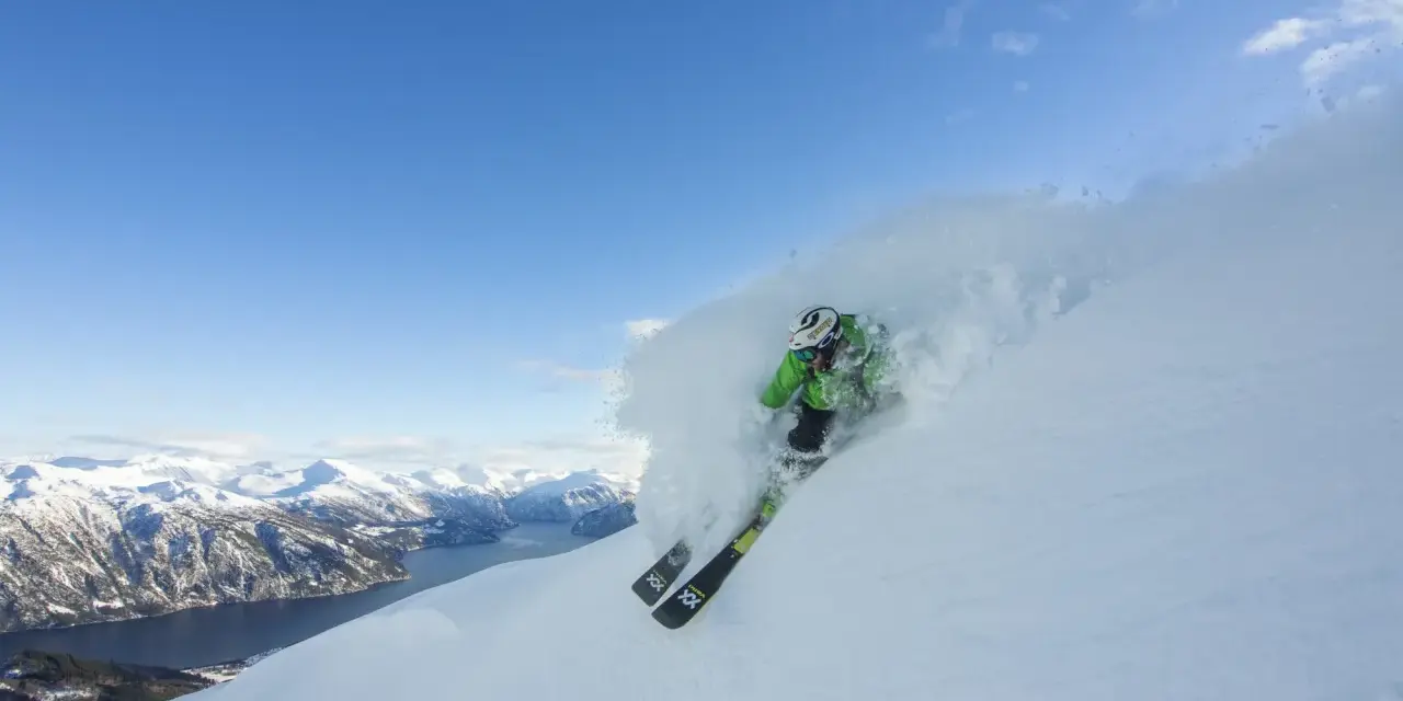 En skidåkare i grön jacka plöjer genom pudersnö med en fantastisk fjordvy i bakgrunden. Stranda ski-skidorna skär genom snön.
