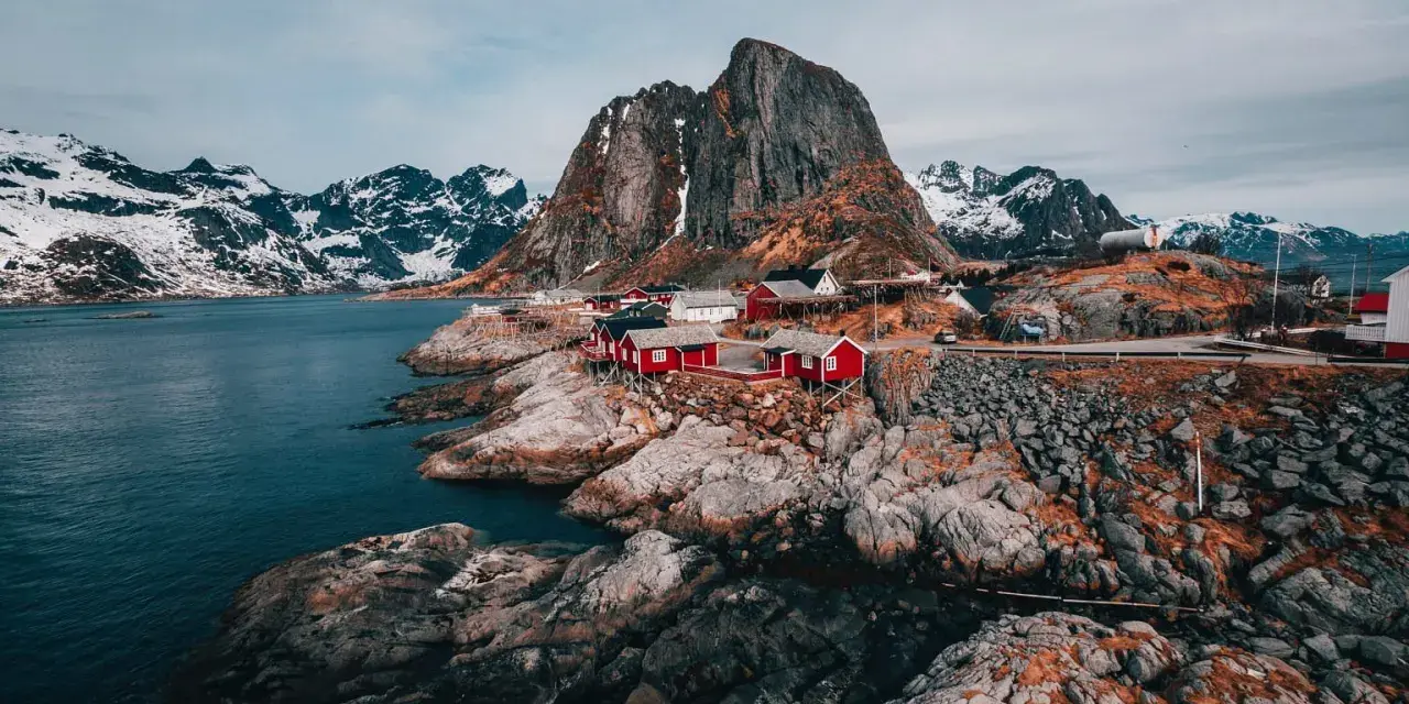 Röda fiskestugor vid en klippig kust i **Hadeland, Norge**, med snöklädda berg i bakgrunden.