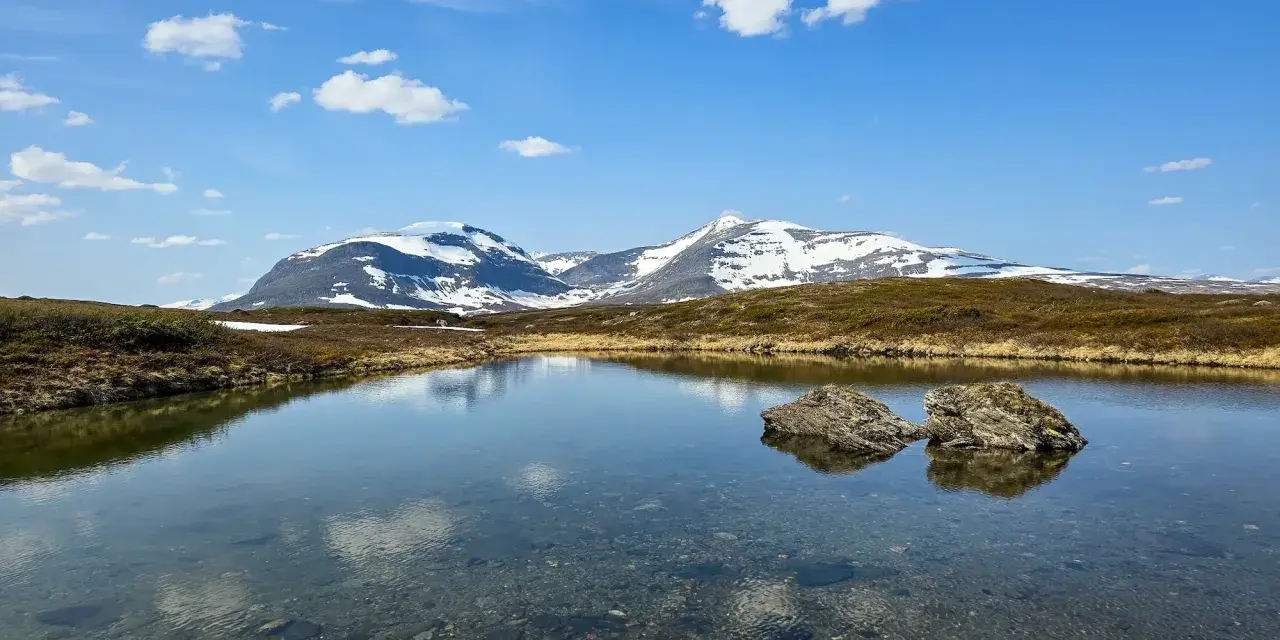 En klarblå himmel med moln speglar sig i en lugn sjö. I bakgrunden reser sig snöklädda berg, en vacker vy för ramundberget vandring.