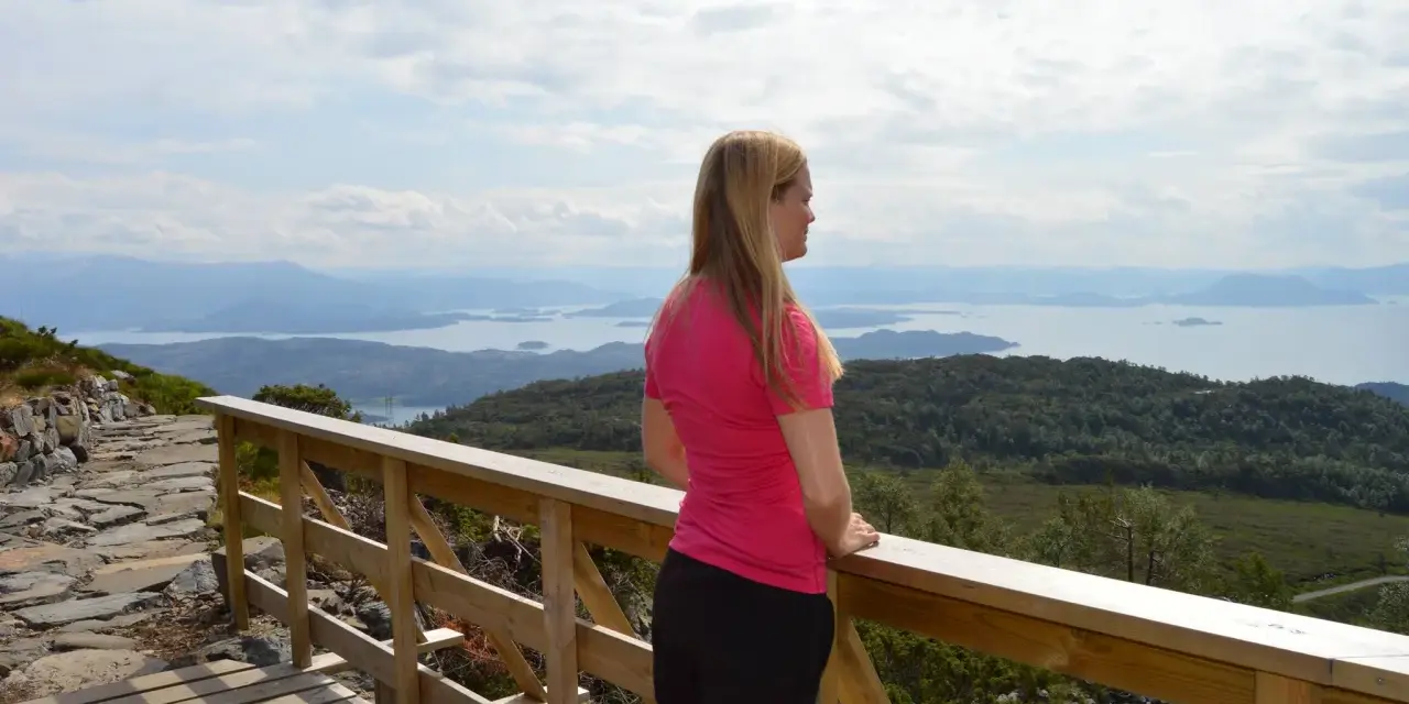 Kvinna i rosa tröja njuter av utsikten över ett storslaget Norge med fjäll och fjordar.