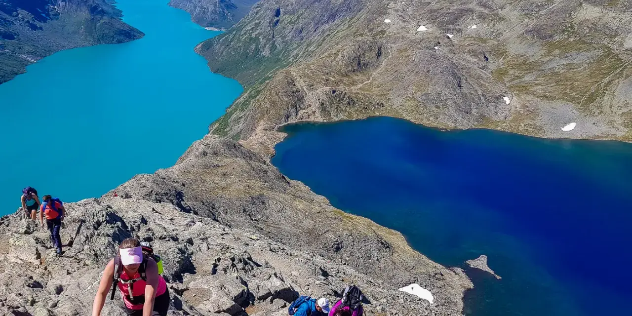 Vandrare klättrar på en stenig bergssida med turkosa och blå sjöar nedanför. En klassisk Besseggen vandring.