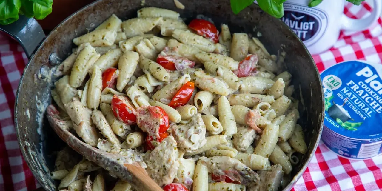 En stor panna med krämig pasta pesto, körsbärstomater och kyckling. En träsked ligger i pannan.