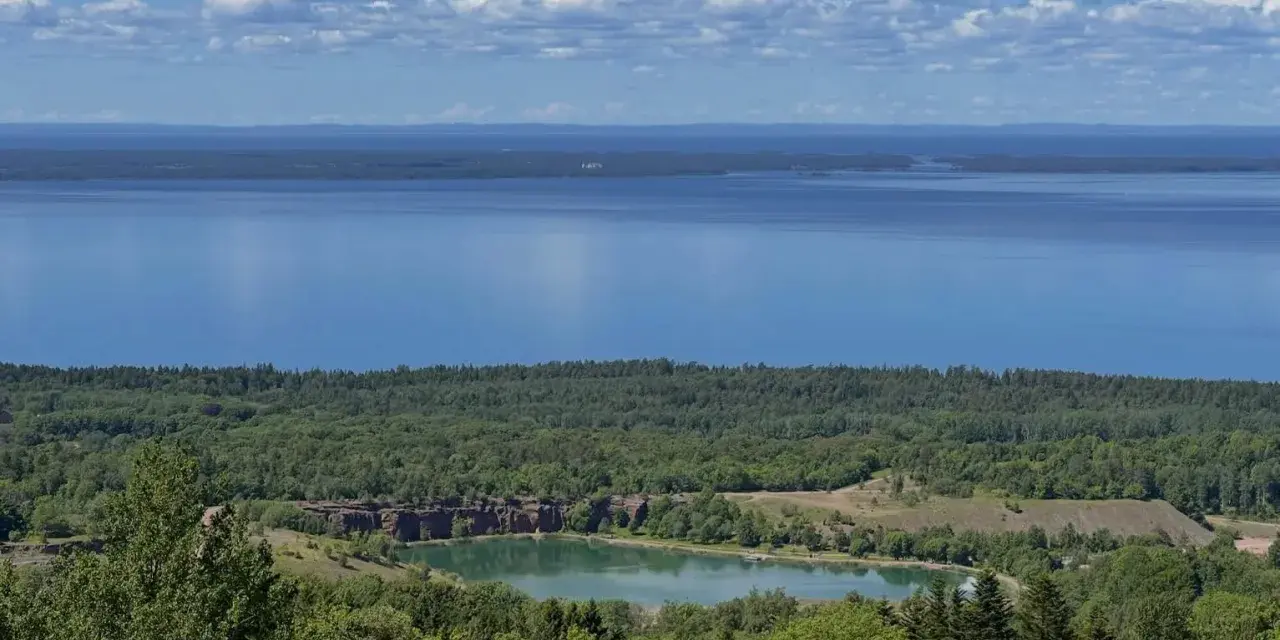 Vacker utsikt över en sjö och en grönskande skog. En liten sjö med klippor syns i mitten, en del av **Götaleden etapp 7**.