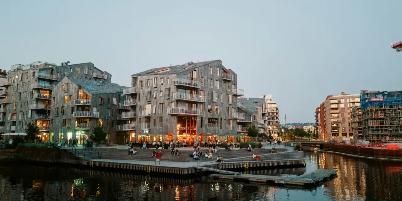 Moderna byggnader längs en kanal i Oslo. Folk sitter på trappor vid vattnet. Besök Oslo för en härlig kväll.
