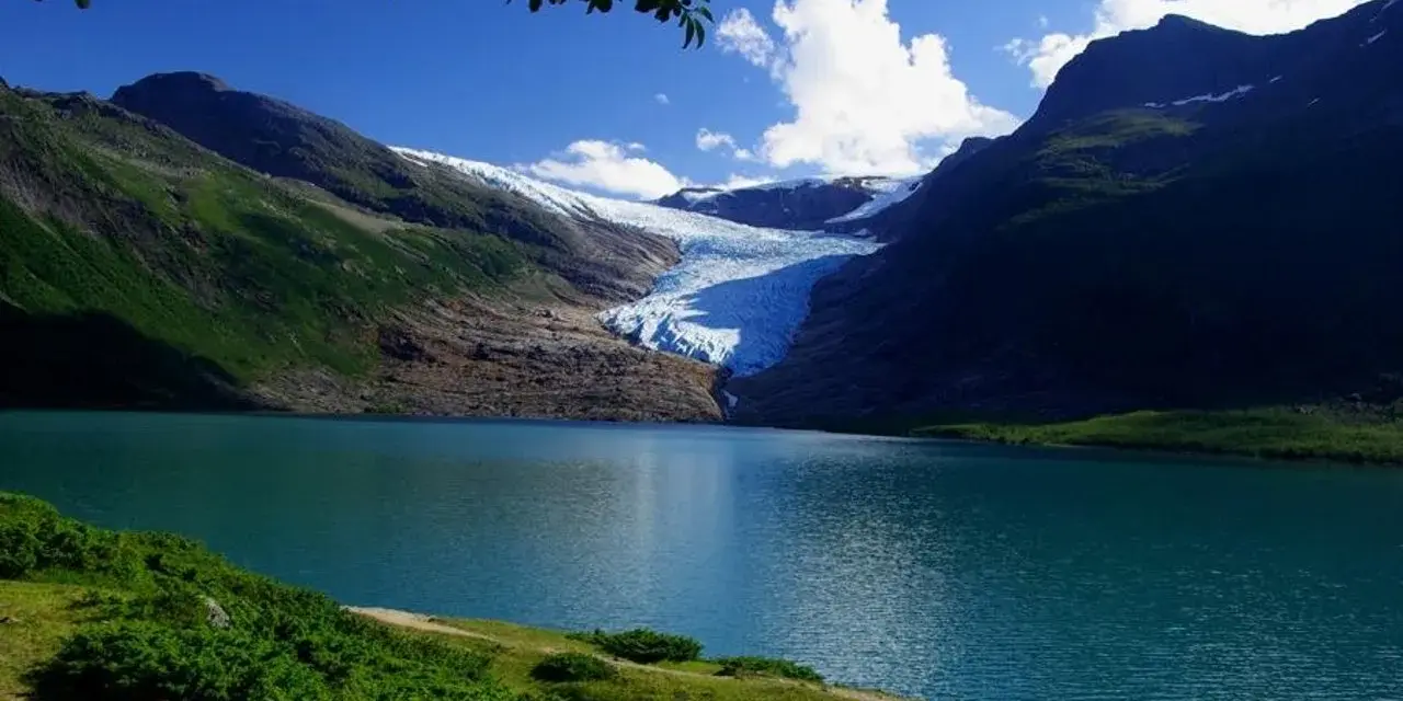 Blå sjö vid foten av en glaciär i Steinkjer, Norge. Grönskande bergssluttningar möter det turkosa vattnet under en klarblå himmel.