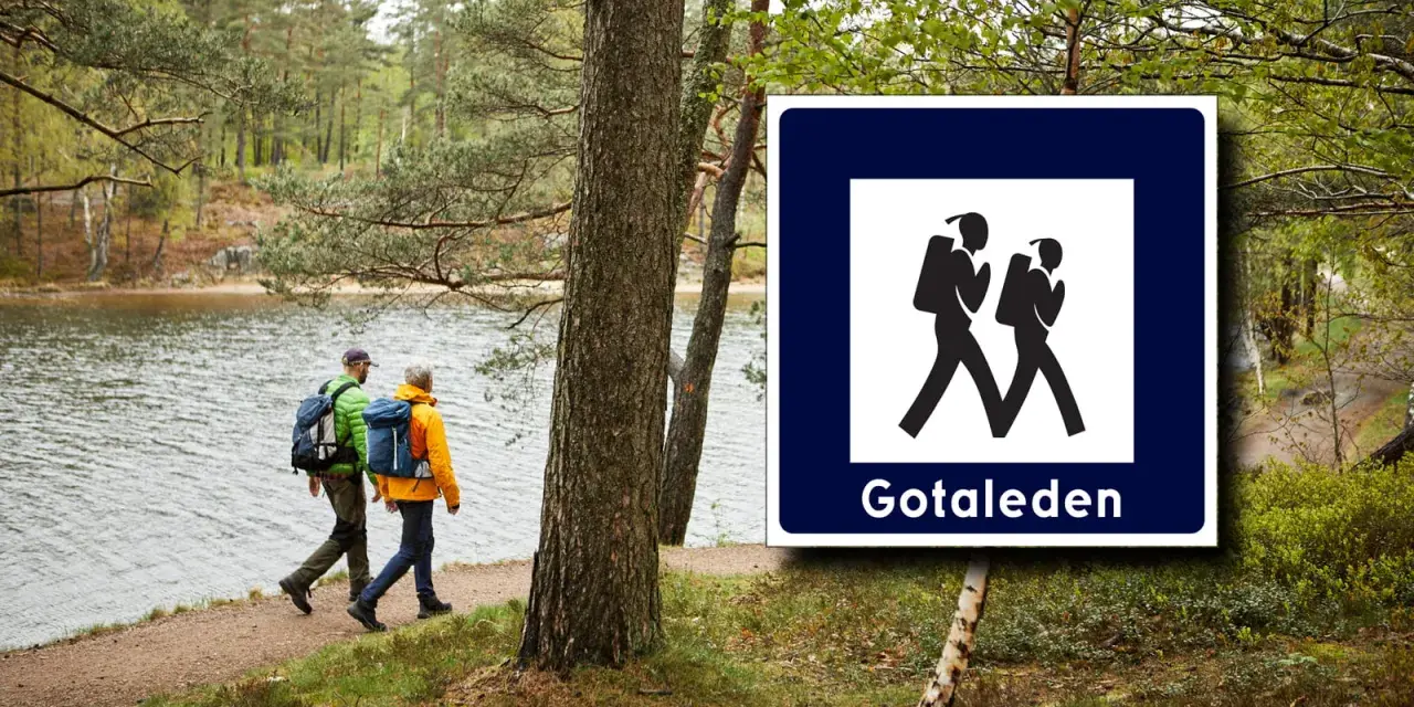 Två vandrare med ryggsäckar följer stigen längs en sjö. Skylt visar vandringsled Gotaleden etapp 6.