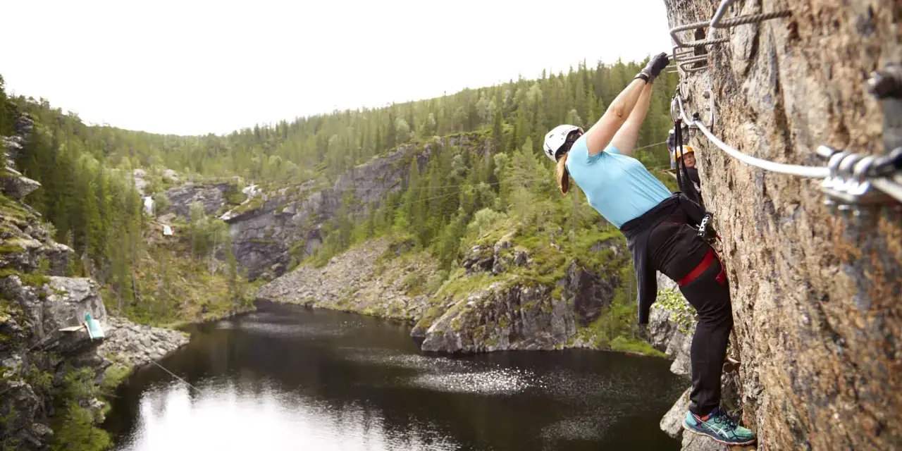 En person klättrar på en klippa vid rypetoppen adventurepark. Vacker natur med sjö och skog.