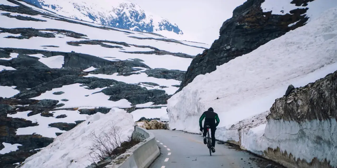 En person cyklar på en snöig väg i Norge, omgiven av höga snödrivor och berg.
