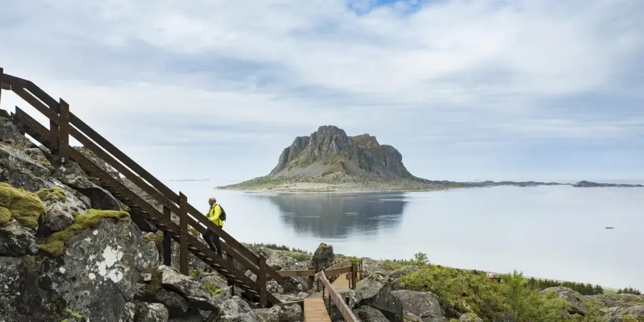 Vandrare på trappor vid havet med en imponerande bergstopp i bakgrunden, en vy från Vega Norge.