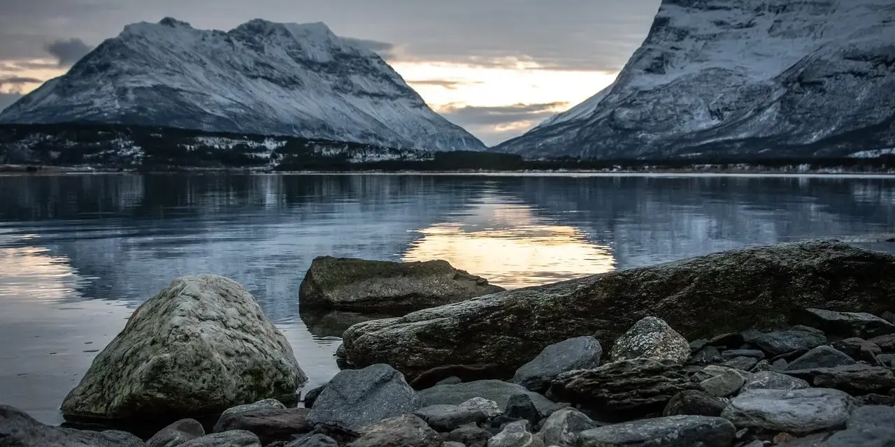 Vinterlandskap med snöklädda berg i Lyngenalperna, speglade i ett lugnt vatten. Stenar ligger vid strandkanten.