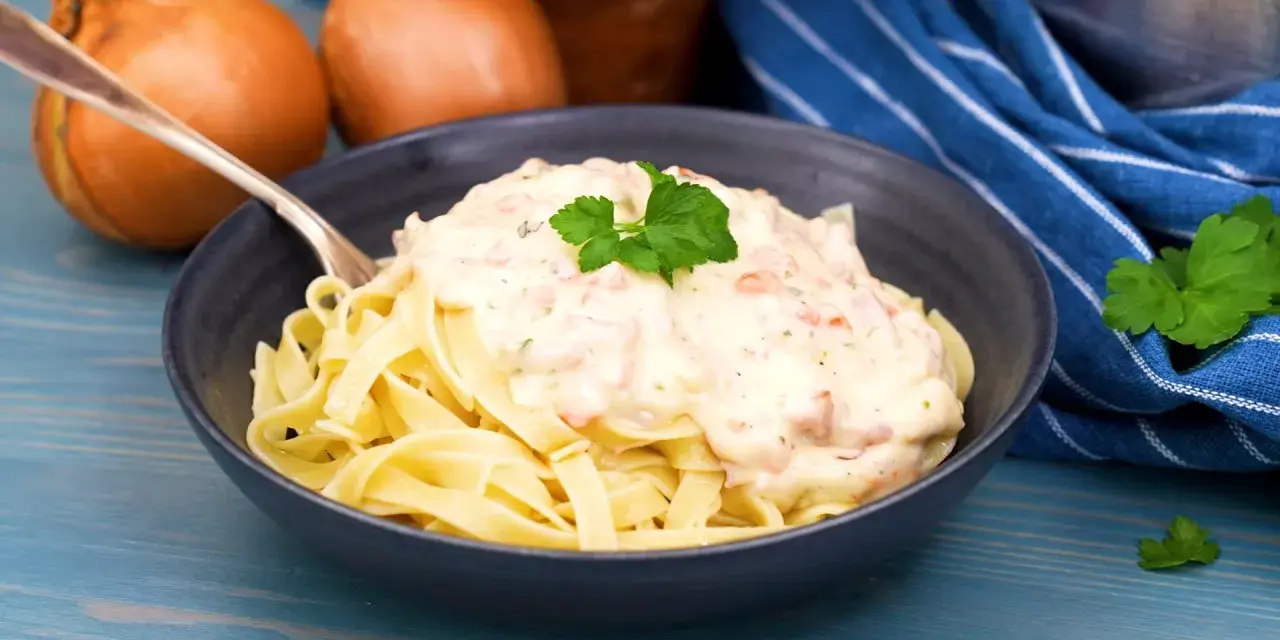 En skål med tagliatelle täckt av krämig ost och skinksås, garnerad med persilja. Två gula lökar syns i bakgrunden.