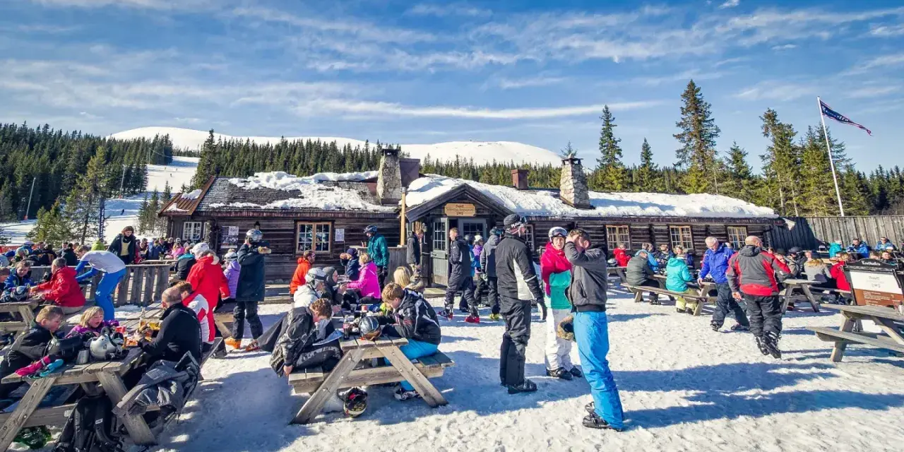 Folk njuter av afterski i Trysil vid en mysig timmerstuga med snöklädda berg och granar i bakgrunden.