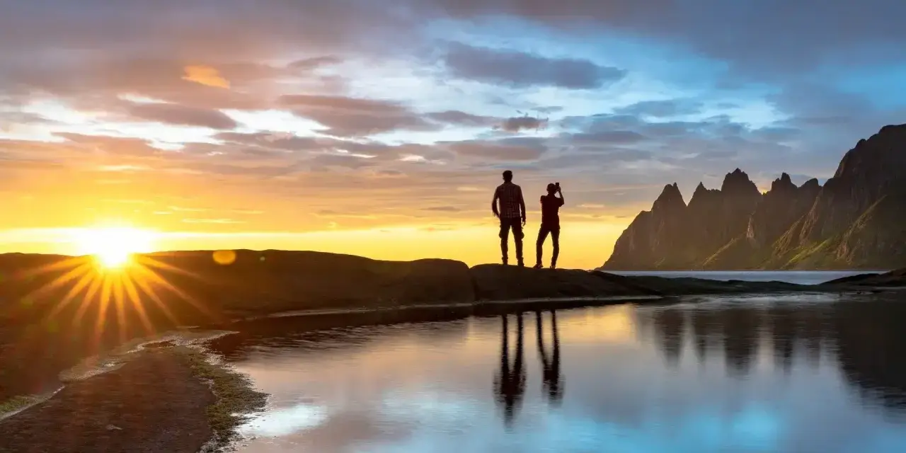 Två silhuetter står på en klippa vid havet och ser på solnedgången över Bodo, Norge. Bergskedja i bakgrunden.