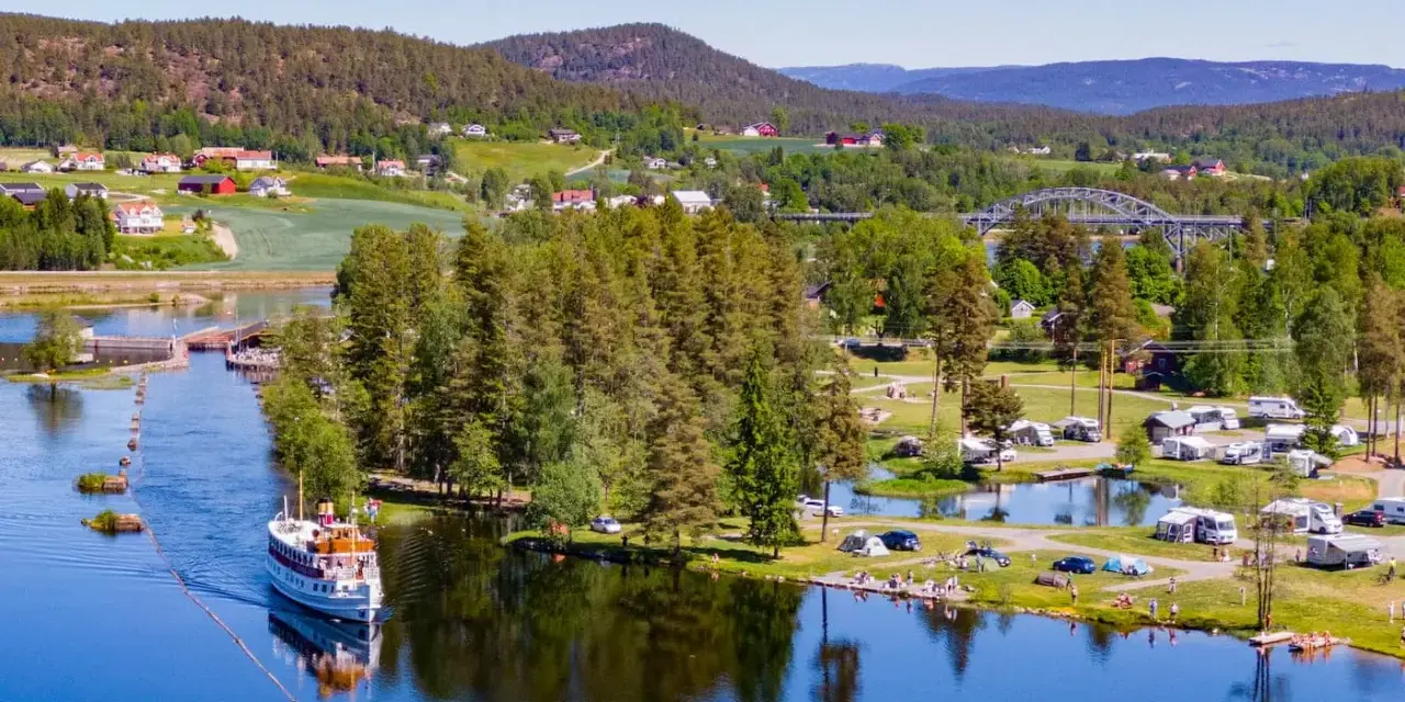 En båt på en sjö vid en campingplats i Norge. Husbilar och tält står utspridda på gräsmattor vid vattnet, omgivna av träd och kullar.