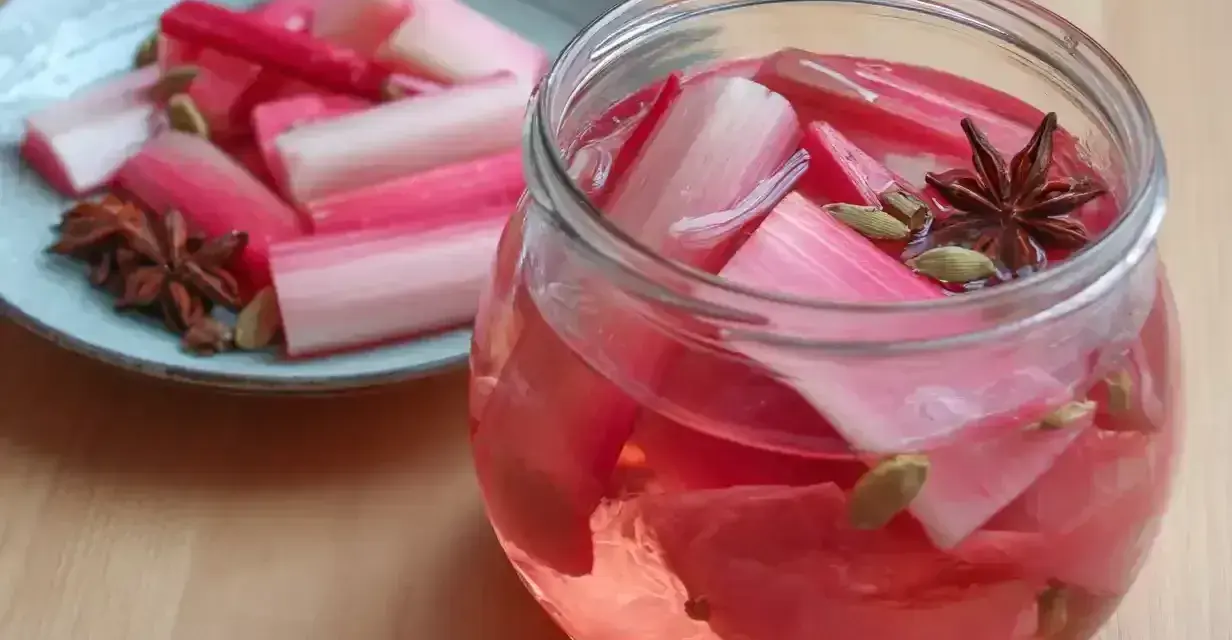 Rabarbersaft recept: färska rabarberbitar i en glasburk med kryddor som stjärnanis och kardemumma. En tallrik med rabarber och kryddor står bredvid.