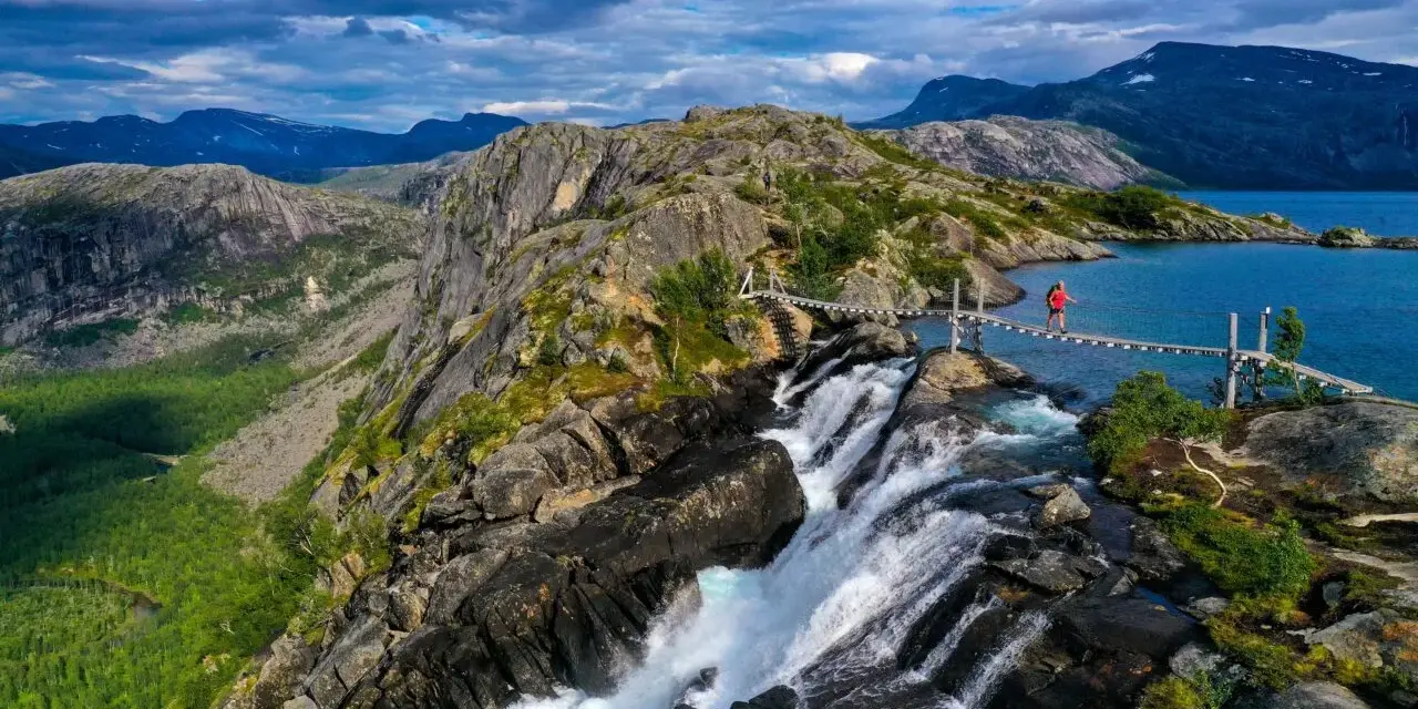En person på en hängbro över ett vattenfall under en vandringsresa i Norge. Dramatiskt landskap med berg och en blå sjö.