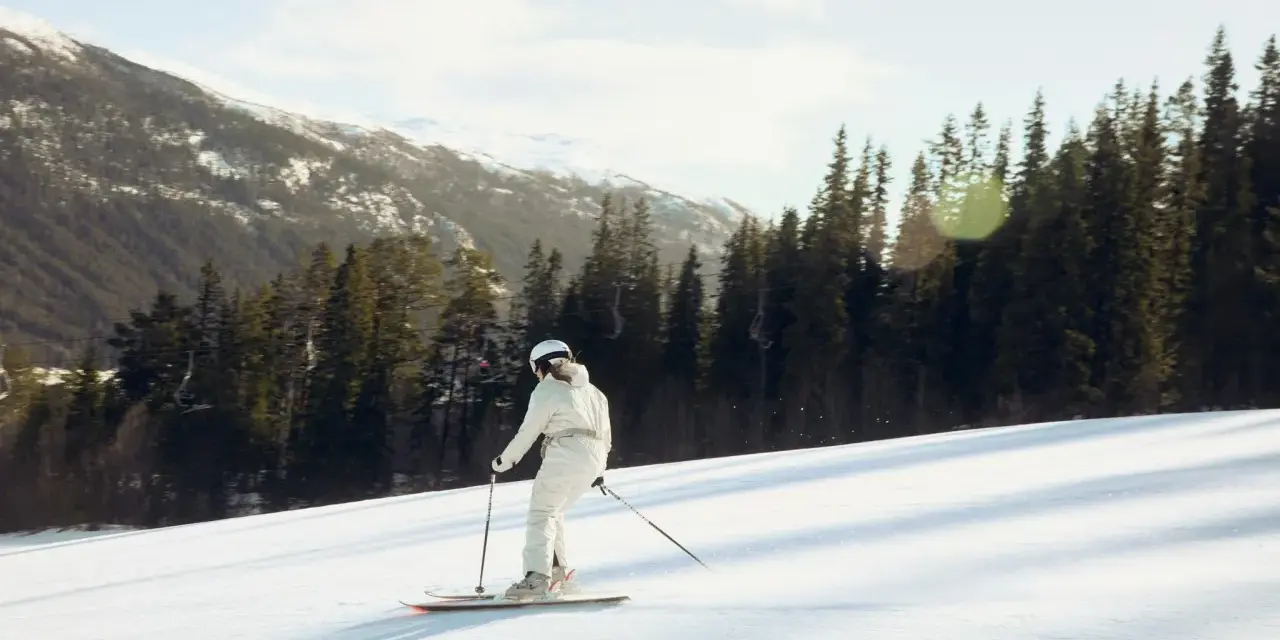 En person åker skidor utför en snöig backe, omgiven av granar och snöklädda berg. En perfekt dag på en av Sveriges skidorter.