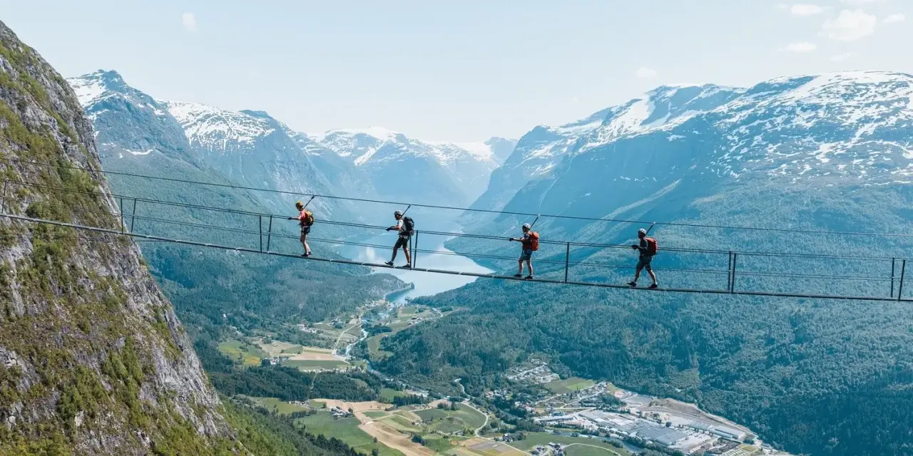 Fyra personer går på en hängbro vid Loen via ferrata, med en fantastisk utsikt över fjord och snöklädda berg.