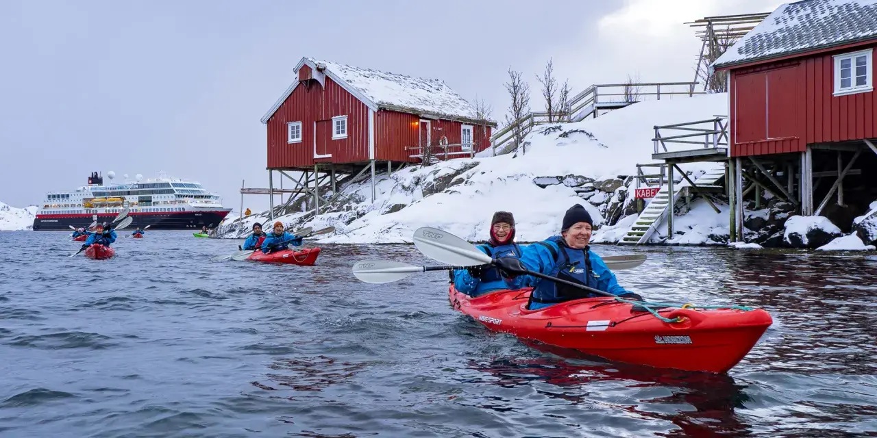 Glada paddlare i kajak i Lofoten, med snöklädda röda hus och ett stort fartyg i bakgrunden.