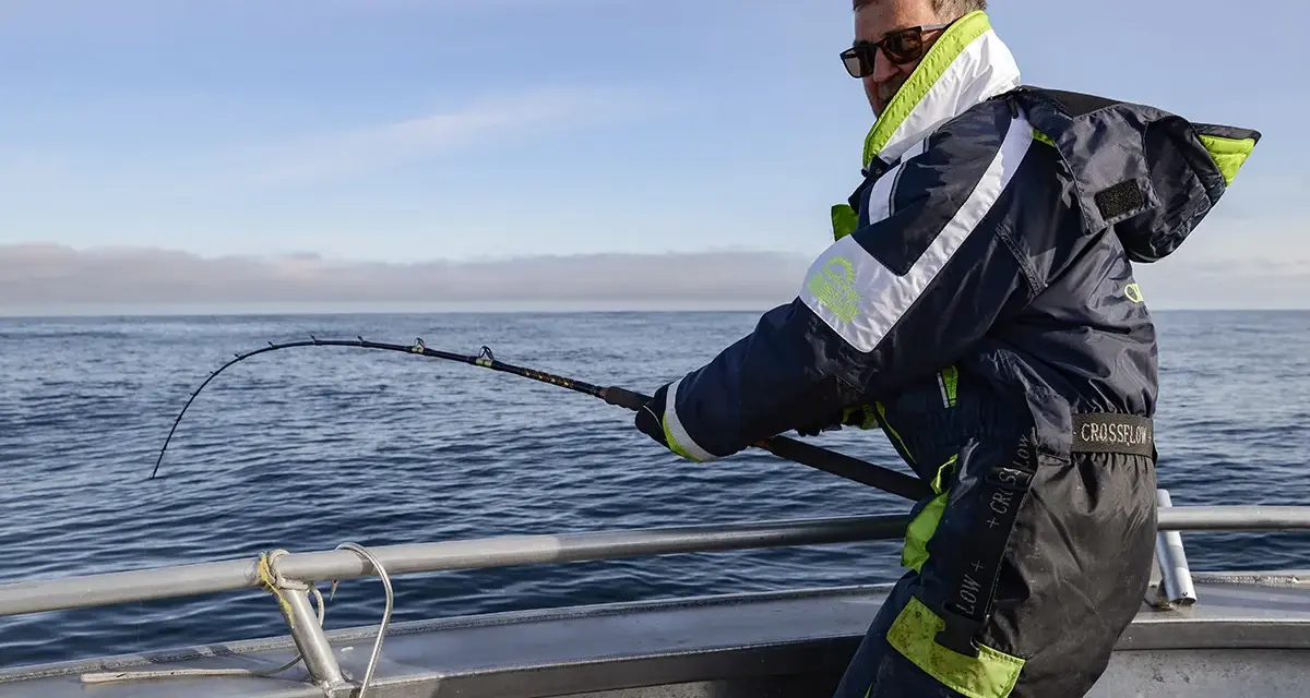Man med solglasögon och fiskeutrustning kämpar med en böjd spö ombord på en båt. En spännande dag med fiska i Norge.
