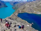 Vandrare klättrar på en stenig bergssida med turkosa och blå sjöar nedanför. En klassisk Besseggen vandring.